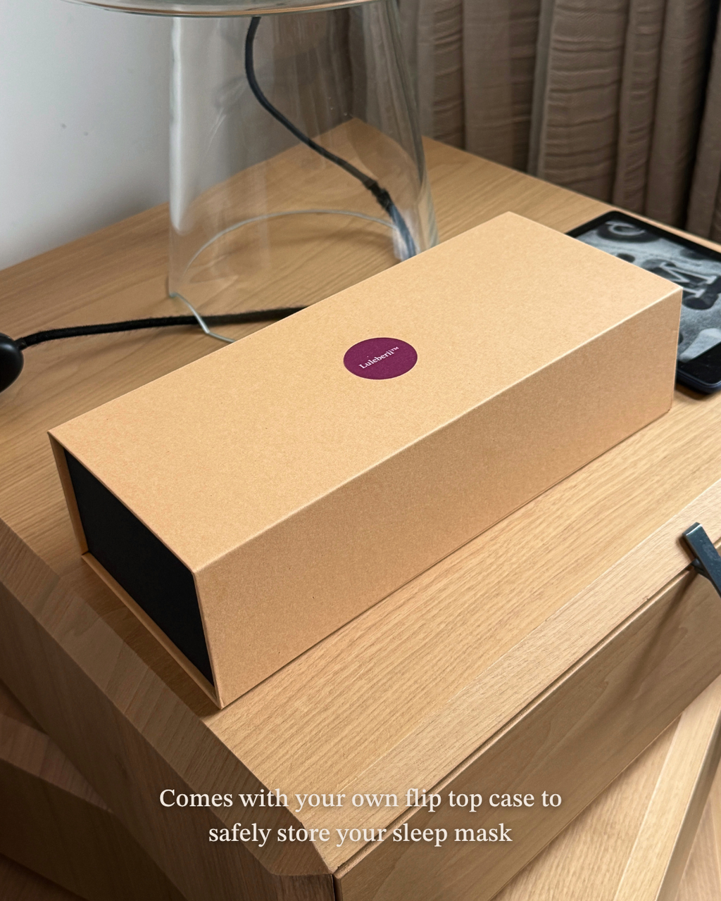 A Kraft colored flip top box with a burgundy circular Luleberii logo sits on a wooden bedside table next to a glass lamp base and a Kindle. Text at the bottom reads, 'Comes with your own flip top box to safely store your sleep mask'