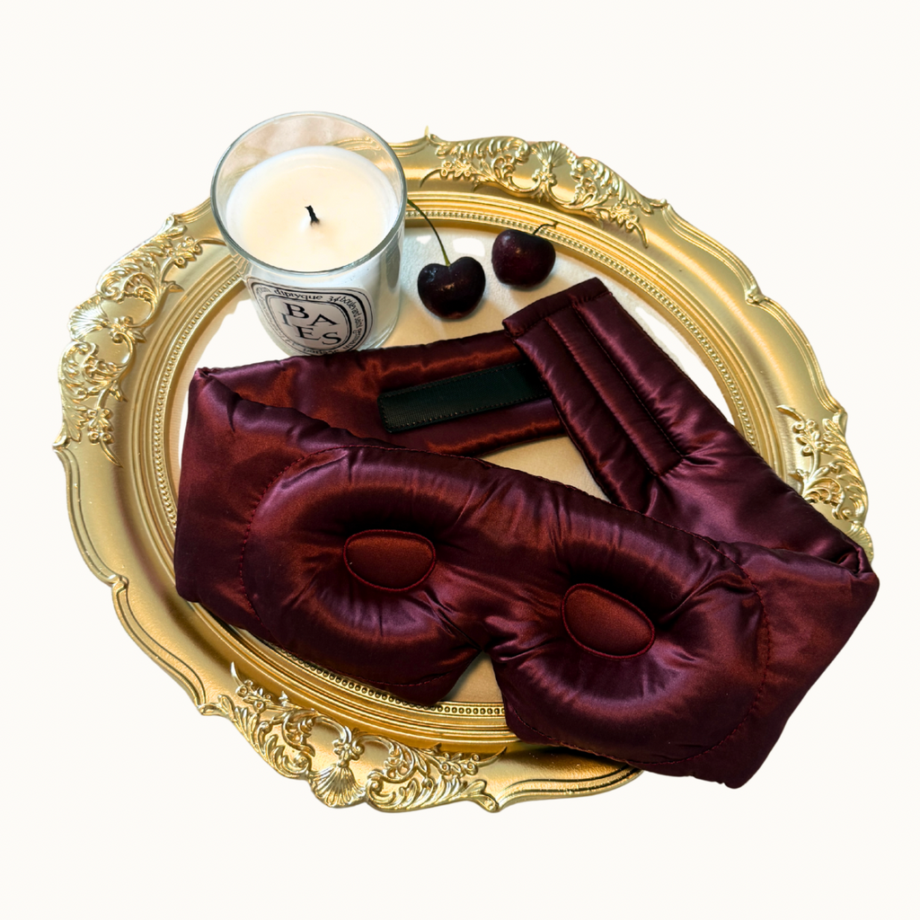 A luxury silk sleep mask in deep burgundy, styled on an ornate gold tray alongside a Parisian-style candle, cherries, and a black rectangular accessory, set against a clean off-white background.