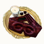 A luxury silk sleep mask in deep burgundy, styled on an ornate gold tray alongside a Parisian-style candle, cherries, and a black rectangular accessory, set against a clean off-white background.