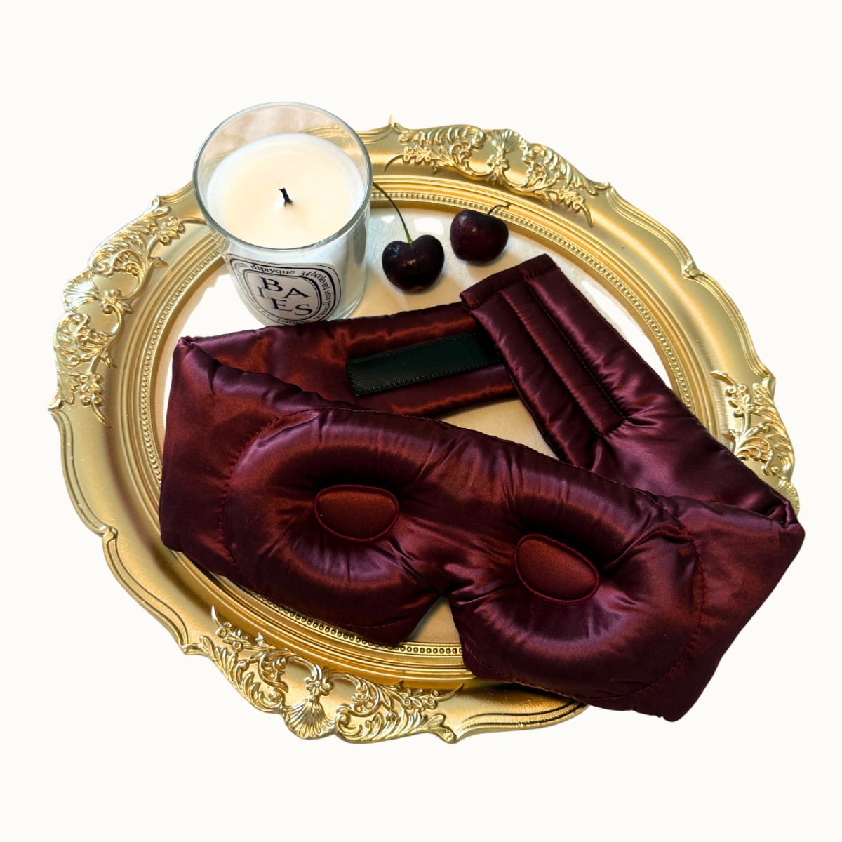A luxury silk sleep mask in deep burgundy, styled on an ornate gold tray alongside a Parisian-style candle, cherries, and a black rectangular accessory, set against a clean off-white background.