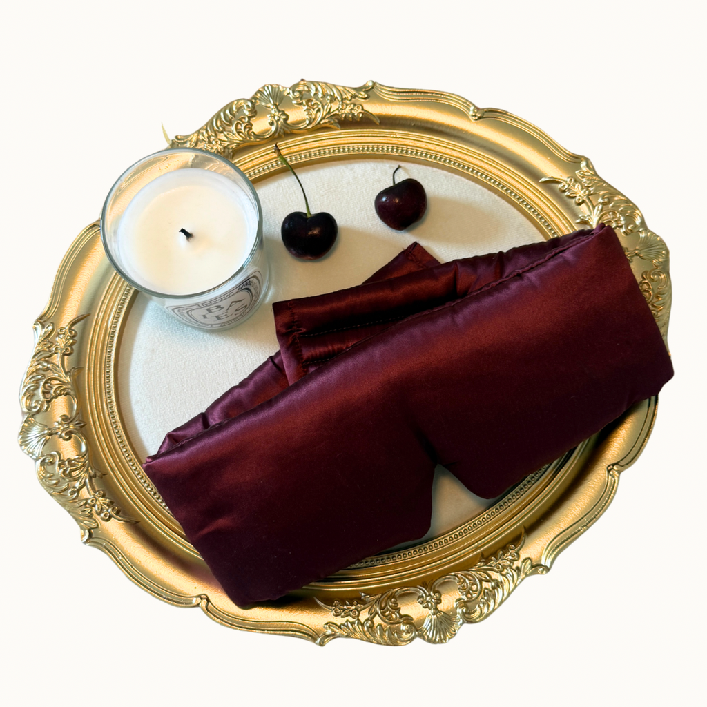 A folded luxurious silk sleep mask arranged on a ornate gold tray with a white candle and two dark cherries on a plain white background.