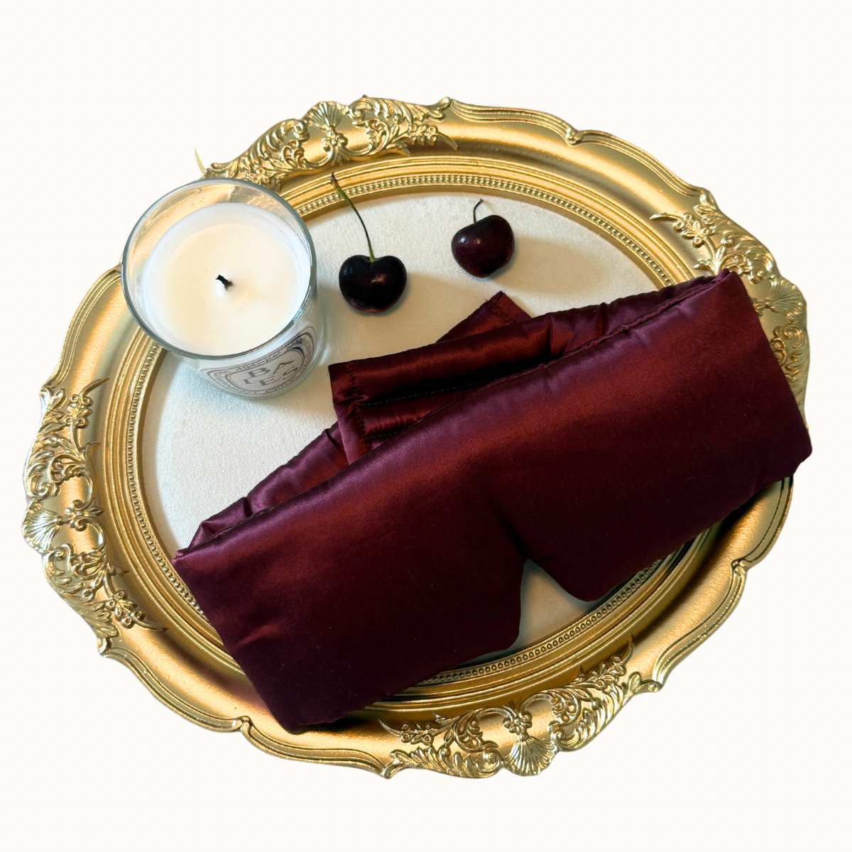 A folded luxurious silk sleep mask arranged on a ornate gold tray with a white candle and two dark cherries on a plain white background.