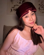 A woman wearing a burgundy pure silk sleep mask on her head, in a light pink eyelet top, posing in a dimly lit indoor setting.